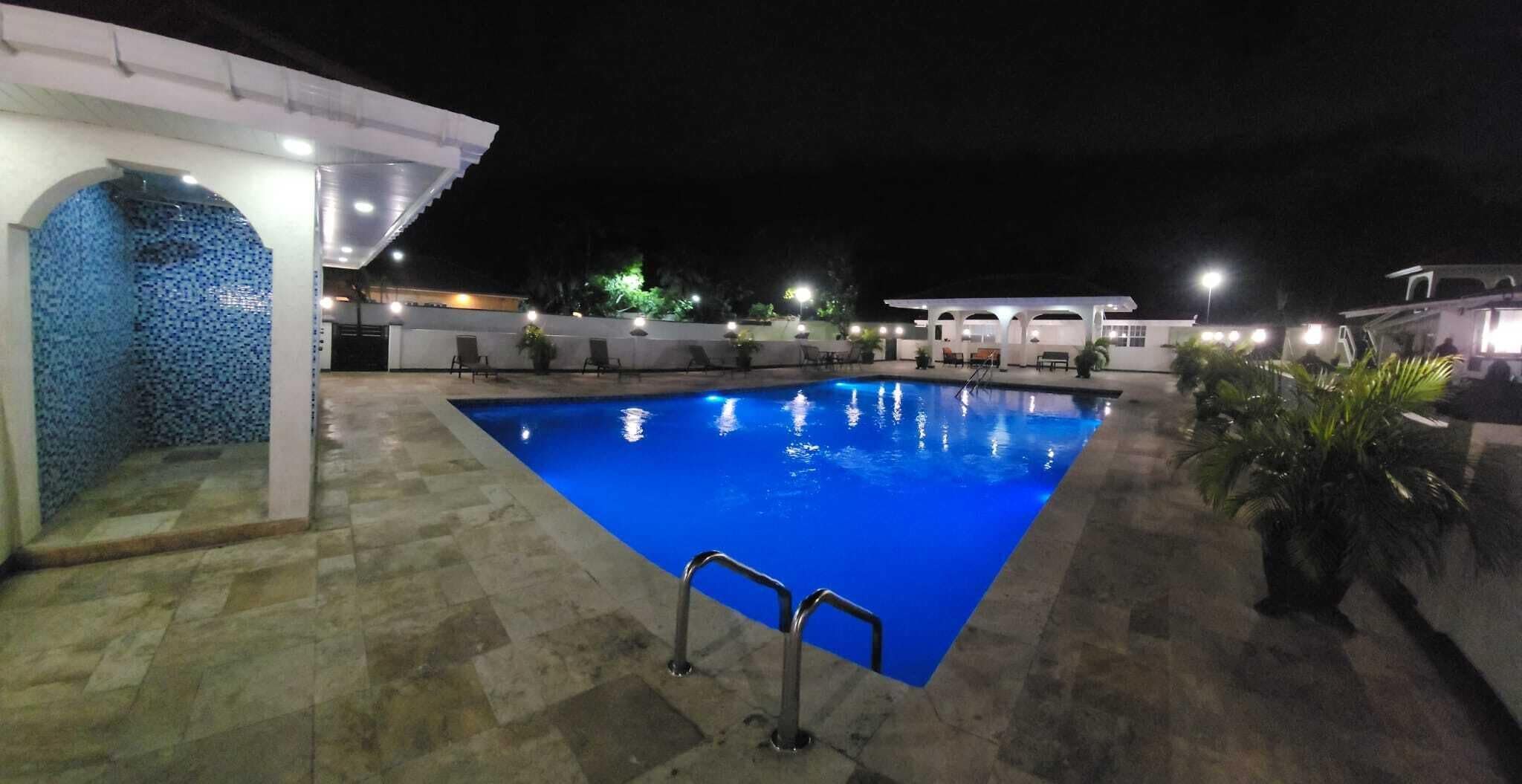 A rectangular swimming pool with a tiled deck surrounding it, a covered seating area with chairs, and a small covered structure with a tiled wall.