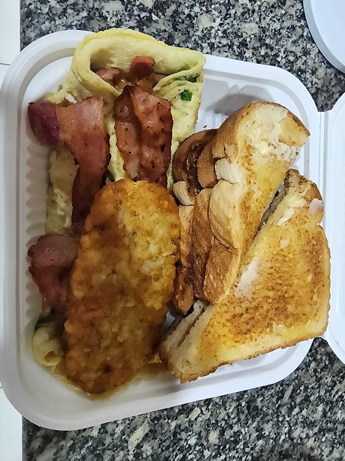 A meal in a takeout container with scrambled eggs, bacon, fried chicken, and toasted bread.