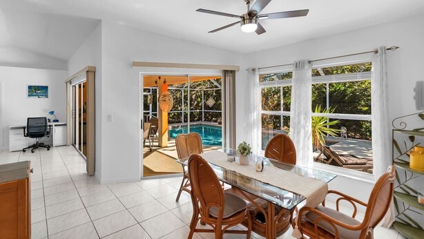 Une cuisine moderne avec des meubles blancs, un coin repas doté d’une table en verre et de chaises en osier, et une vue sur une piscine à travers de grandes fenêtres.