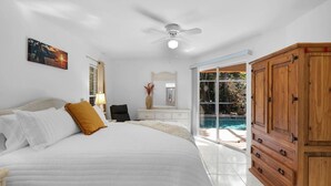 Une chambre à coucher comprenant un lit, un ventilateur de plafond, une table de chevet, une lampe, un miroir, une armoire en bois et une porte-fenêtre coulissante donnant sur une piscine.