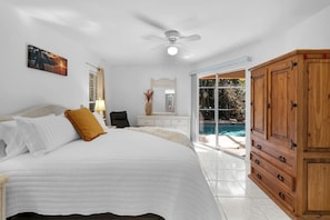 Una camera da letto con un letto, una ventola a soffitto, un comodino, una lampada, uno specchio, un'armadio in legno e una porta a vetri scorrevoli che dà su una piscina.
