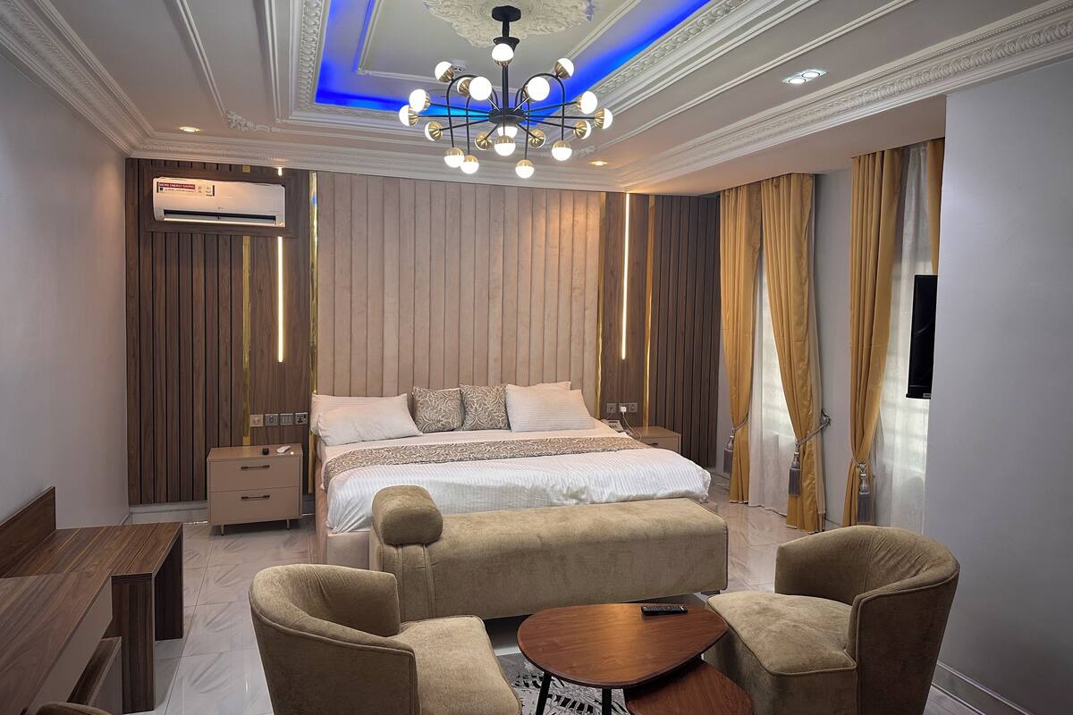 A modern bedroom with a large bed, a chandelier, a wooden panel wall, and a seating area with a table and chairs.