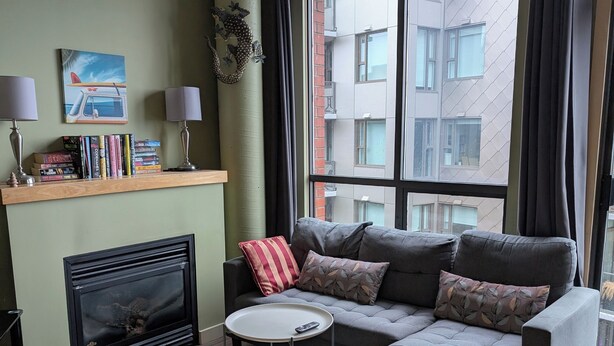 Ein modernes Wohnzimmer mit einem grauen Sofa, einem Kamin, einem Bücherregal und einem großen Fenster mit Vorhängen.