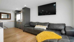 A modern living room with a sofa, a TV mounted on the wall, and a kitchen area in the background. - Sleeps 16 - 15 min/Downtown, 10 min/Medical Center (Aurora)