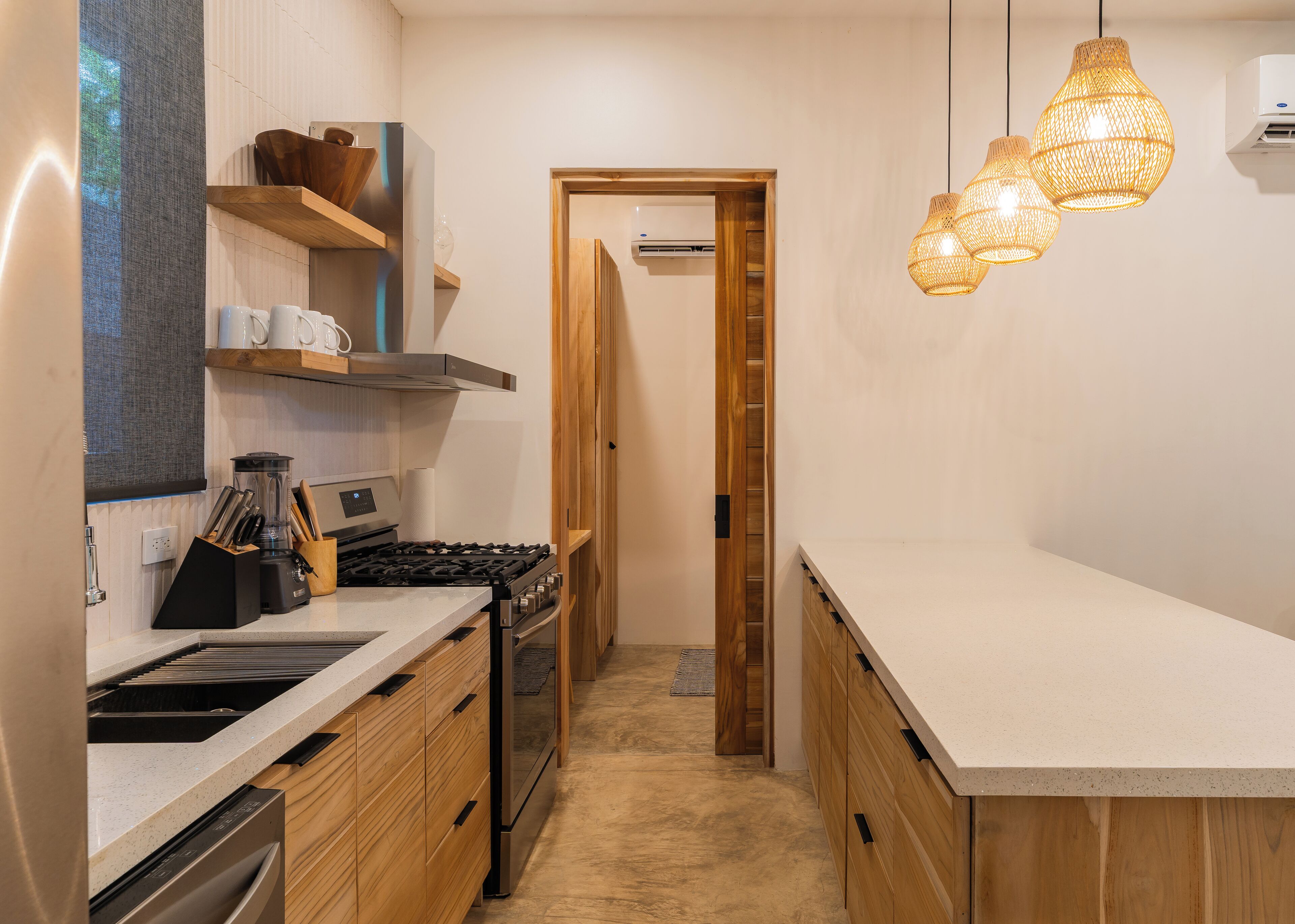 Une cuisine étroite avec des meubles en bois, une cuisinière en acier inoxydable et un plan de travail. On y trouve des luminaires suspendus en osier et une porte qui donne sur une autre pièce.