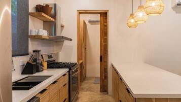 Une cuisine étroite avec des meubles en bois, une cuisinière en acier inoxydable et un plan de travail. On y trouve des luminaires suspendus en osier et une porte qui donne sur une autre pièce.