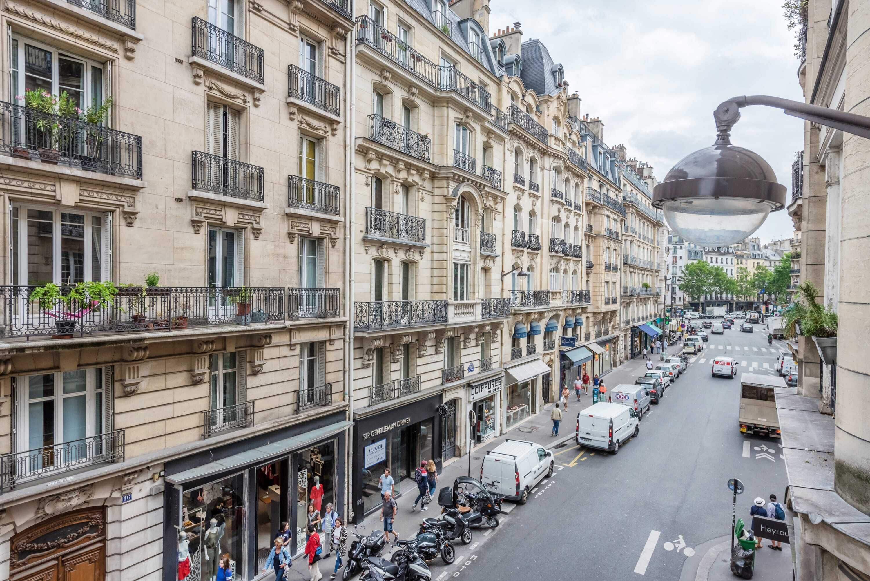 A street view with multi-story buildings, parked vehicles, and pedestrians.