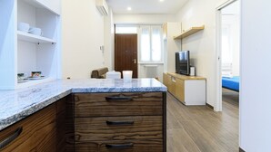 Una cocina compacta con encimera de mármol, armarios de madera, televisión empotrada y una pequeña zona de estar con una silla y una mesa.