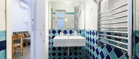 Een badkamer met een witte wastafel, blauw-witte tegels, een handdoekrek en een stoel in de aangrenzende kamer.