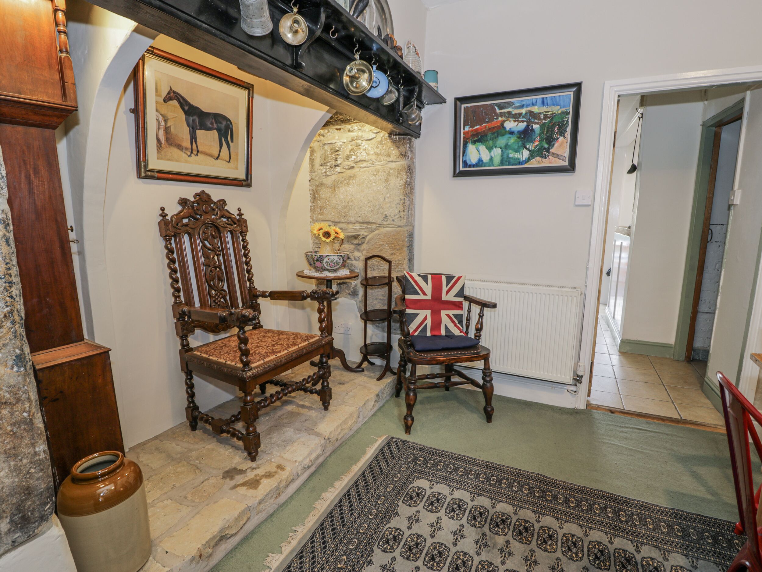 Una habitación tradicional con chimenea de piedra, una silla de madera con cojín con la bandera Union Jack, una silla de madera antigua y un cuadro enmarco de caballos.