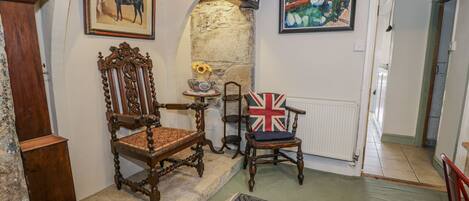 Una habitación tradicional con chimenea de piedra, una silla de madera con cojín con la bandera Union Jack, una silla de madera antigua y un cuadro enmarco de caballos.