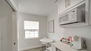 Una cocina moderna con armarios blancos, microondas y cafetera. Hay una ventana con persianas, una mesita redonda y una silla.