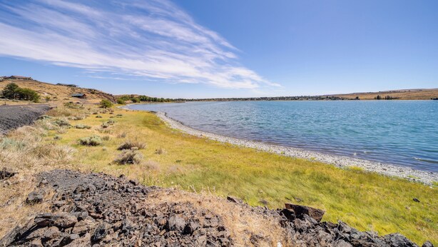 Blick auf einen See mit steiniger Uferlinie, grünen Flächen und sanften Hügeln im Hintergrund.
