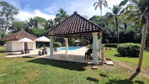 Un pabellón con techo de tejas, rodeado por una piscina, palmeras y un área verde con mobiliario exterior.