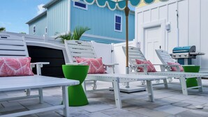 A patio area with white lounge chairs and green stools, a grill, and a blue building in the background. - 4BR beach club w/ Hot tub + Pool + Game room (North Myrtle Beach)