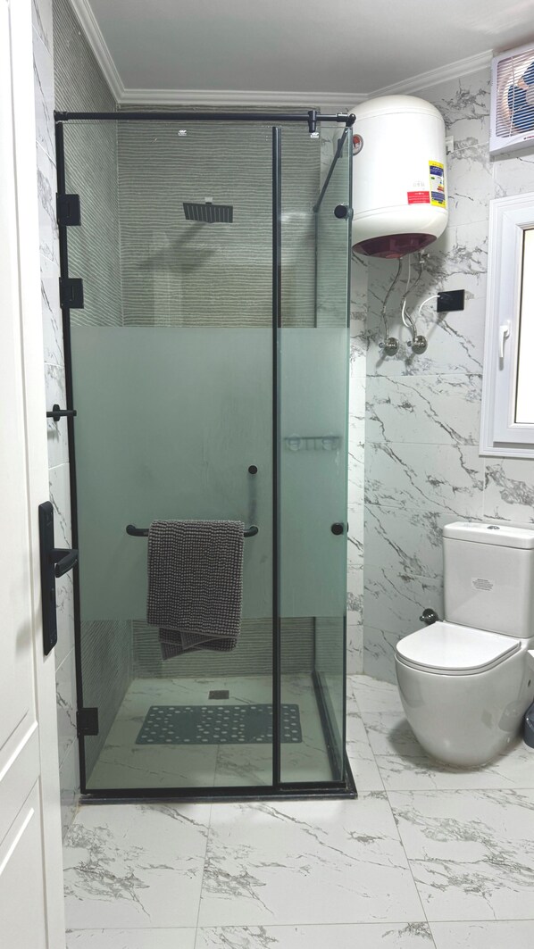 A glass-enclosed shower stall with a towel rack, a toilet, and a water heater in a bathroom with marble flooring.