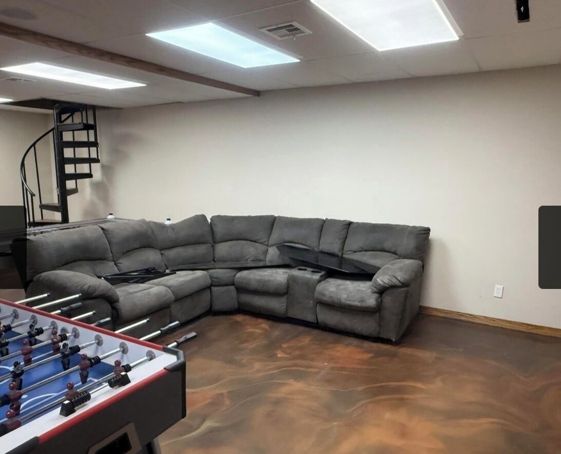 A room with a gray sectional sofa, a foosball table, and a spiral staircase.