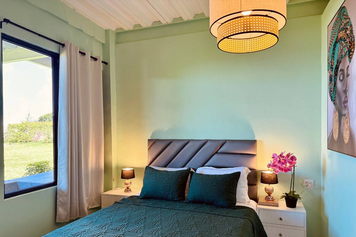 A bedroom with a bed, nightstands, a lamp, a window with curtains, and a wall with a painting.