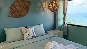 A bed with a white comforter and two pillows, a woven wall hanging, a hanging light fixture, and a window with a view of a landscape. - Lake View House Cerro Verde (Santa Ana)