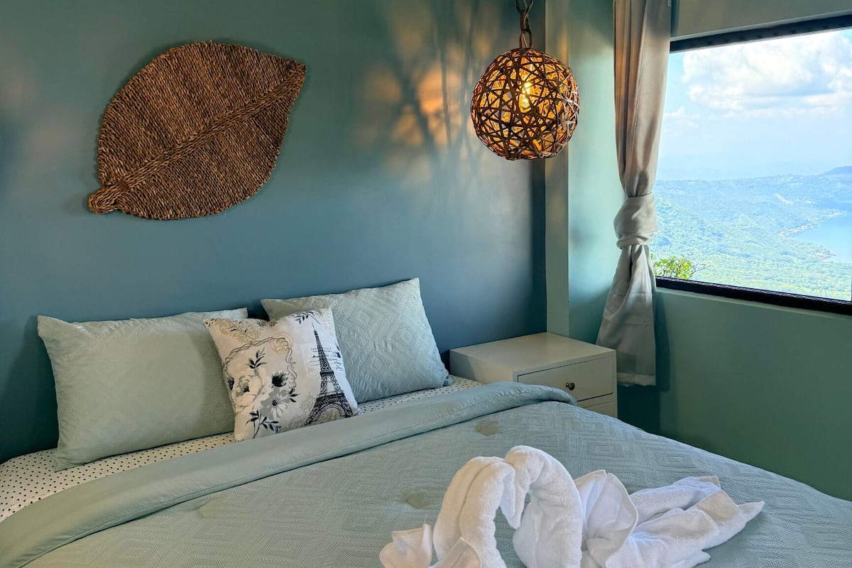 A bed with a white comforter and two pillows, a woven wall hanging, a hanging light fixture, and a window with a view of a landscape.
