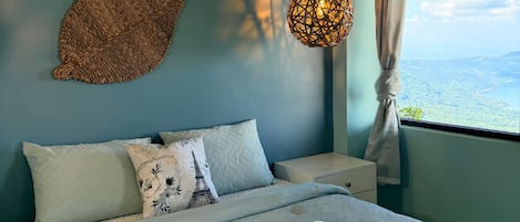 Una cama con edredón blanco y dos almohadas, un tapiz de pared tejido, una lámpara colgante y una ventana con vistas al paisaje.