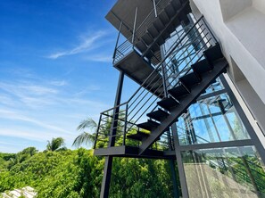 A modern staircase with glass railings, leading to a building with a clear sky and lush greenery in the background. - 2BR at Rizz Suites • Pool • 24/7 GYM • Walk to Beach • 2 King Beds • Sosua (Sosúa)