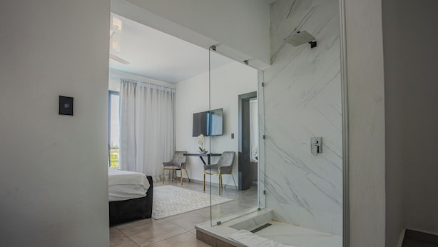 A modern hotel room with a glass-enclosed shower, a bed, a desk with a chair, and a TV mounted on the wall.