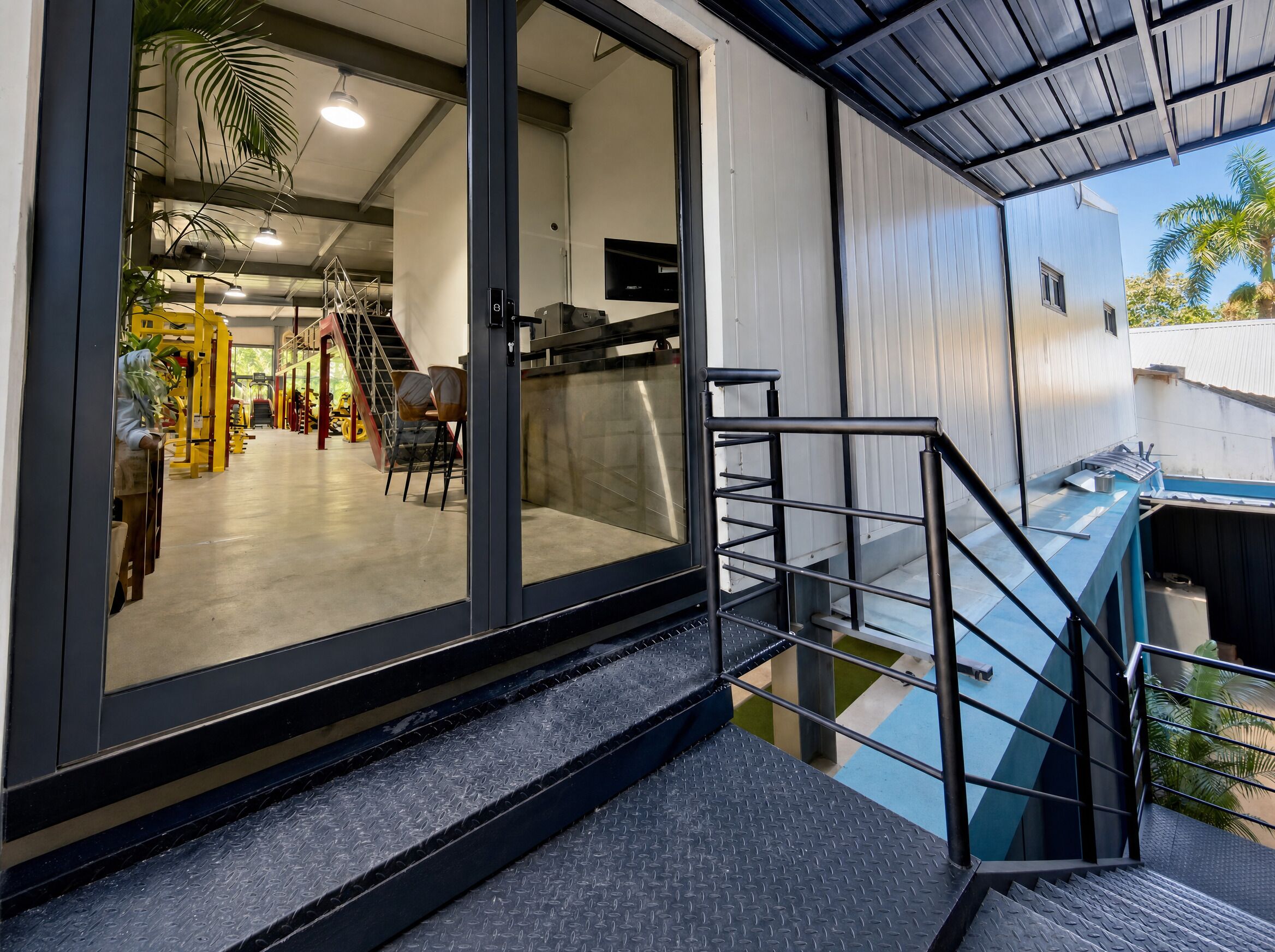 Un interior moderno con una escalera que conduce a un balcón, una entrada con puerta de vidrio y un espacio exterior visible con palmeras.