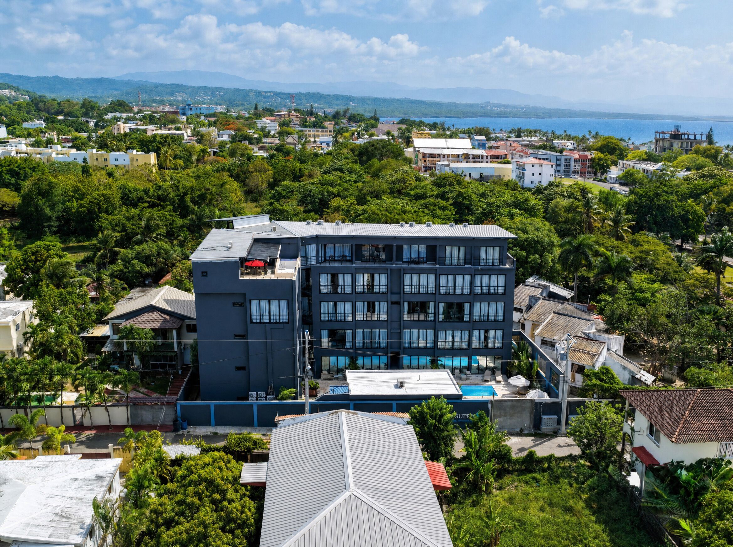 A multi-story hotel with a rooftop pool, surrounded by lush greenery and a view of the sea.