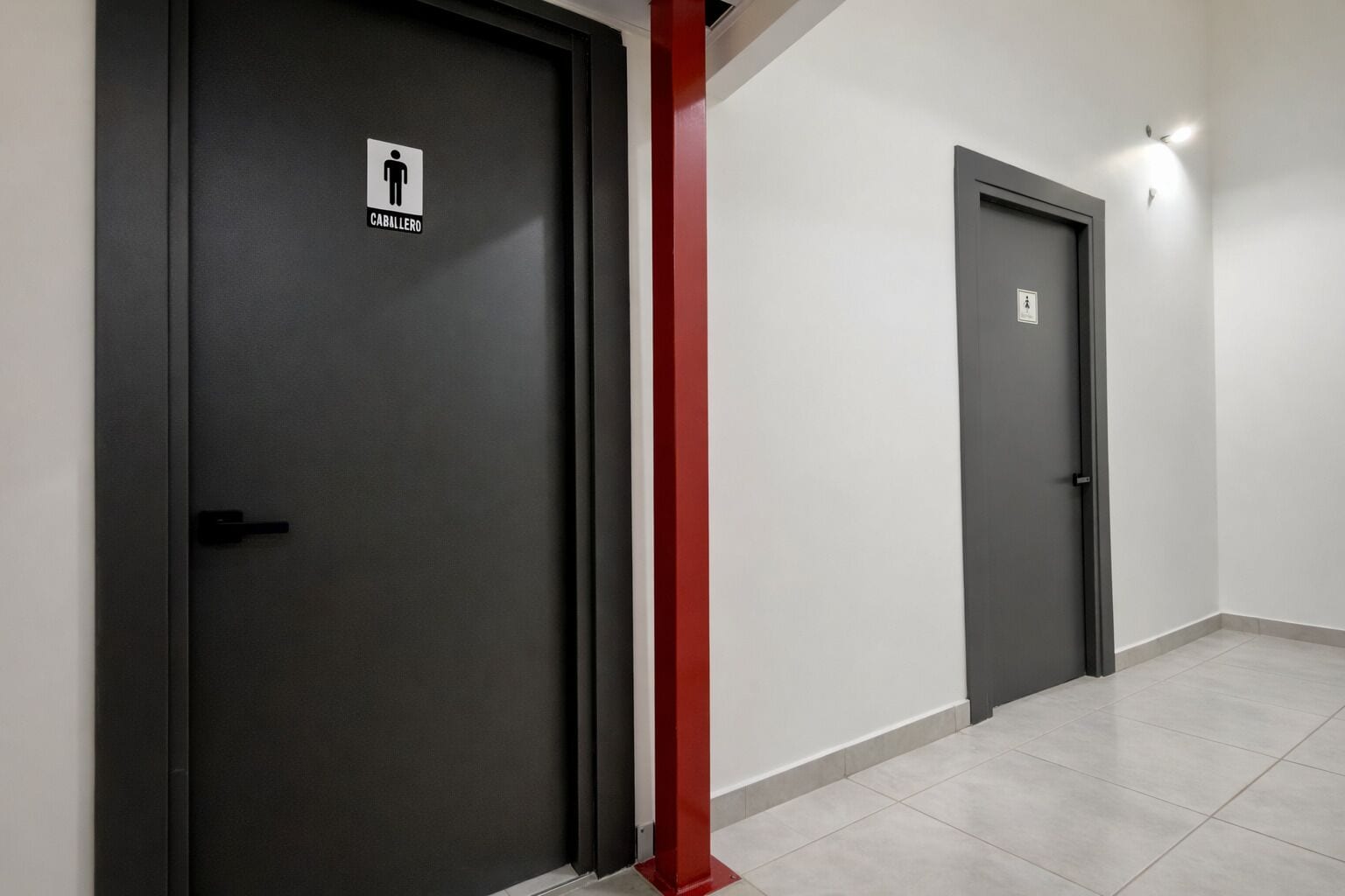 To mørke døre med symbol for herretoilet, en rød bæredygtig søjle og et flisegulv.