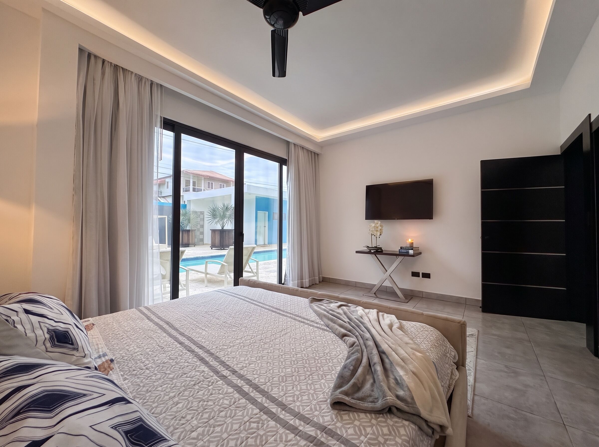 Ein modernes Hotelzimmer mit einem großen Bett, einem Flachbildfernseher, einem Schreibtisch mit Lampe und Blick auf einen Swimmingpool.