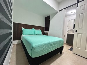 Un lit simple avec une couverture matelassée, une tête de lit sombre, une porte blanche et une petite table de chevet.
