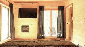 A wooden cabin room with a bed, a window with curtains, and a small fireplace. - Peaceful Pines (South Bloomingville)