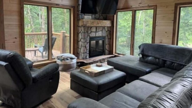 A cozy cabin interior with a stone fireplace, leather sofas, and large windows offering a view of the surrounding forest. - Peaceful Pines (South Bloomingville)