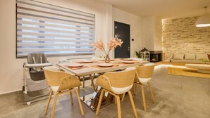 Een moderne eetruimte met een grote tafel, zes stoelen en een kinderstoel. Er is een groot raam met jaloezieën, een stenen muur en een open haard.