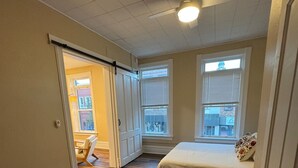 A bedroom with a bed, a chair, and a ceiling fan. There are two windows with blinds and a door leading to another room.