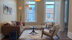 A living room with a sofa, two armchairs, a coffee table, and a rug. There are two windows with views of buildings outside.