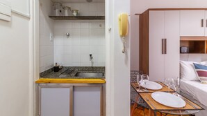 Une petite cuisine équipée avec un évier et un plan de travail, un coin repas avec une table pour deux personnes, et un lit avec un oreiller et une couverture.