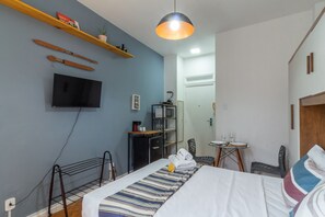 Ein Hotelzimmer mit Bett, Wandfernseher, einer kleinen Küchenzeile, einer Essecke mit Tisch und Stühlen sowie einer Deckenleuchte.