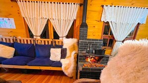 A cozy cabin interior with a wooden floor, a blue sofa, a fur-covered chair, and a fireplace with logs.
