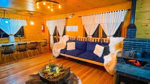 A cozy cabin interior with a wooden floor, a sofa, a dining table with chairs, a fireplace, and a hanging light fixture.