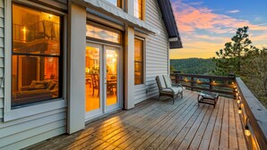 Una terraza de madera con mobiliario exterior, una entrada con puerta de cristal y una vista panorámica de un área boscosa.