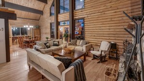 Interior de una cabaña de troncos con un amplio salón, pisos de madera y un comedor visible a través de un vidrio.