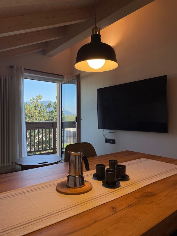 A dining area with a wooden table, a coffee pot, and several cups. A TV is mounted on the wall, and a window with a view of trees and mountains is visible. - La Téza Penthouse (Trentino-Alto Adige)