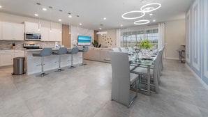 A modern kitchen with a central island, bar stools, a dining area with a glass table, and a large window with a view. - New Jungle Adventure 9BR Villa by Disney (Davenport)