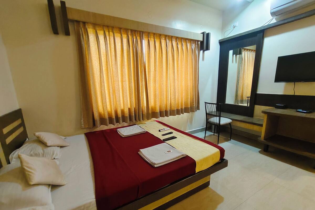 A neatly made bed with a red bedspread and pillows, a bedside table with a remote control, and a television on a stand in a hotel room.