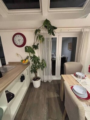 A dining area with a wooden table set for two, a potted plant, a clock on the wall, and a sliding glass door leading to another room. - Chic Riverside Retreat Salford 4bd + free parking (Salford)