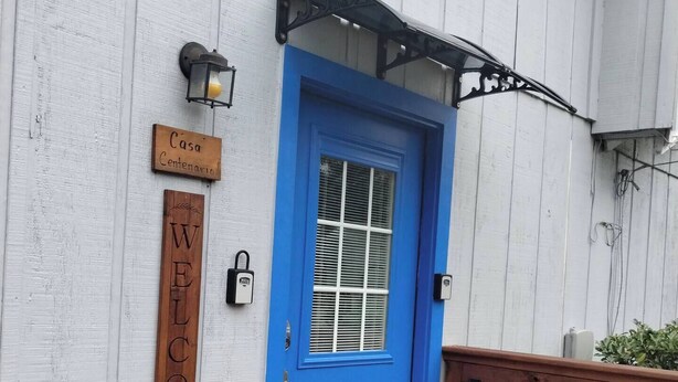 Een blauwe deur met een raam, een 'Welcome' bord en een 'Casa Centenario' bord op een wit houten gebouw.