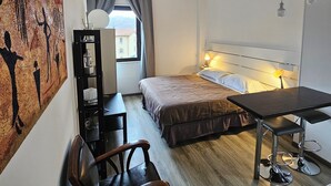 Et lille hotelværelse med seng, skrivebord, stol og bord. Der er et vindue med gardiner, en væglampe og et maleri på væggen.