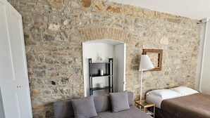 A room with a stone wall, a grey sofa, a bed, a bedside table with a lamp, and a small shelf with a bottle.